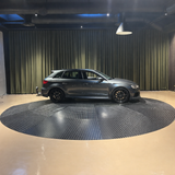 Grå Audi RS3 Sportback set fra siden på rund drejescene i et mørkt bilstudio med gardinbaggrund og loftsbelysning.